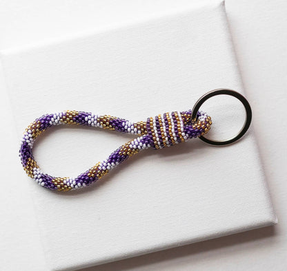 A handmade beaded keychain with a purple, white, and gold patterned loop and a black metal key ring, resting on a white textured surface.