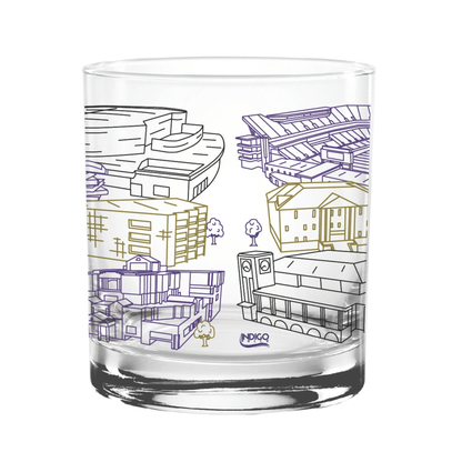 A clear glass rocks tumbler featuring hand-drawn architectural illustrations of various buildings in purple, gold, and black outlines, with a small 'INDIGO' logo at the bottom.