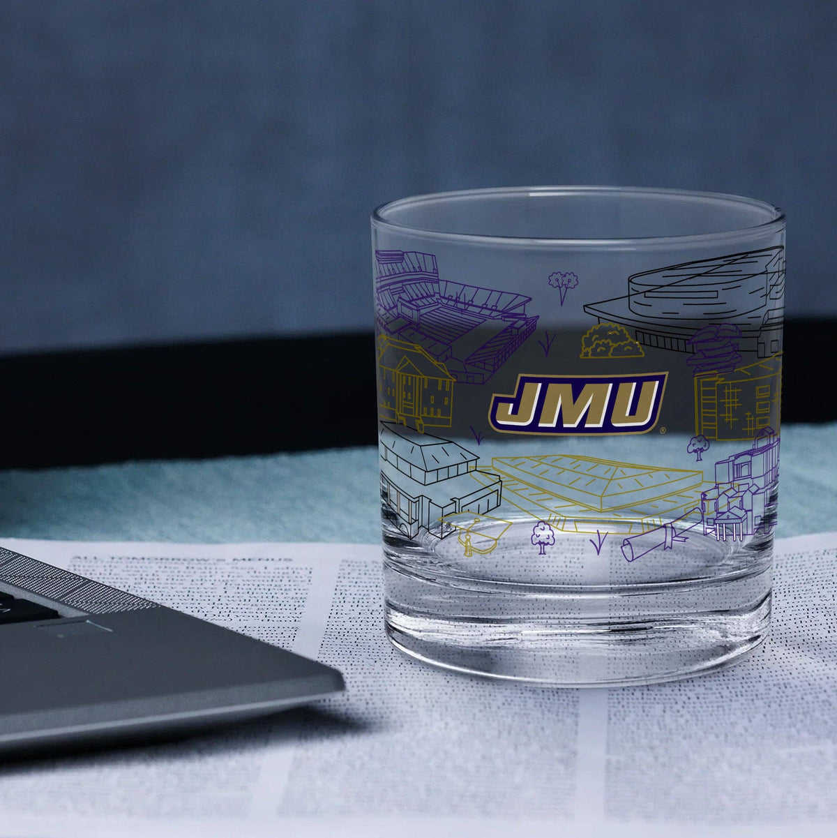 A clear glass tumbler with purple and yellow architectural line drawings and a bold 'JMU' logo, shown on top of an open book next to a laptop in a blue-toned setting.