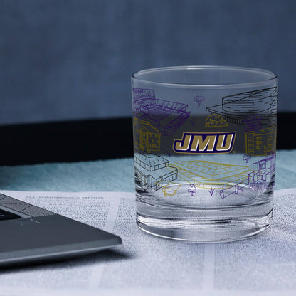 A clear glass tumbler with purple and yellow architectural line drawings and a bold 'JMU' logo, shown on top of an open book next to a laptop in a blue-toned setting.