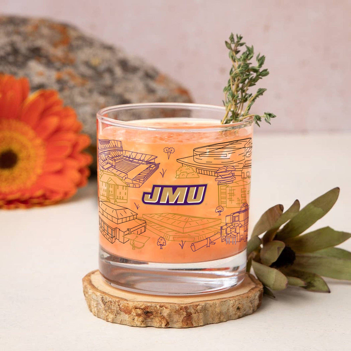 A clear rocks glass with an orange graphic print featuring the letters 'JMU' and architectural sketches, filled with an orange drink and garnished with a sprig of herbs. The glass sits on a round wood slice coaster, with a blurred orange flower and green leaves in the background.