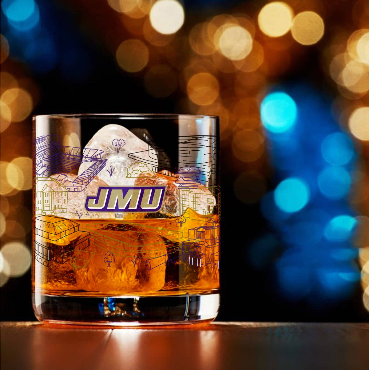 A clear rocks glass filled with amber liquor and large ice cubes, featuring blue line-art campus-style graphics and the letters 'JMU' in purple and white. The glass sits on a dark tabletop with a blurred warm and blue bokeh background.