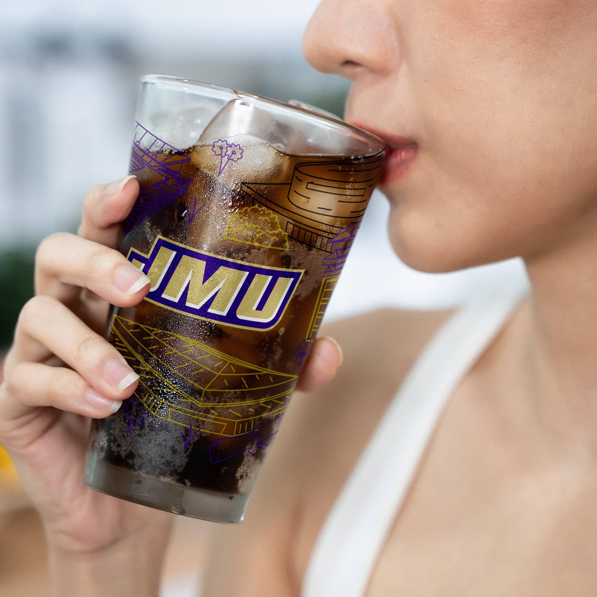 A close-up lifestyle shot of a person drinking from a clear glass tumbler with a dark cola beverage. The glass features a purple and gold graphic design with the letters 'JMU' in white.