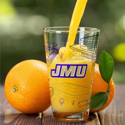 A clear glass tumbler with purple and yellow line drawings and the bold text 'JMU' on the front, filled with orange juice as it is being poured. Two whole oranges with green leaves sit on a wooden surface around the glass, with a blurred green outdoor background.