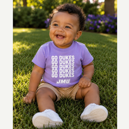 A smiling baby sitting on green grass outdoors, wearing a lavender-purple t-shirt with 'GO DUKES' printed in white text and tan shorts.