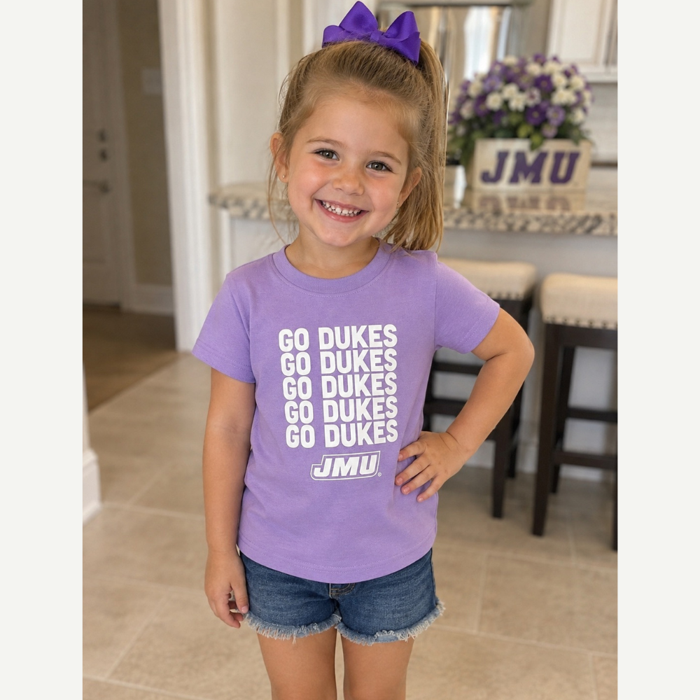 A young girl with a purple hair bow wearing a lavender purple t-shirt with 'GO DUKES' printed in white, paired with denim shorts, standing in a home interior.