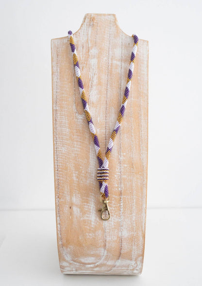 A braided purple, white, and gold lanyard with a gold metal lobster clasp, displayed on a rustic, whitewashed wooden stand against a plain white background.