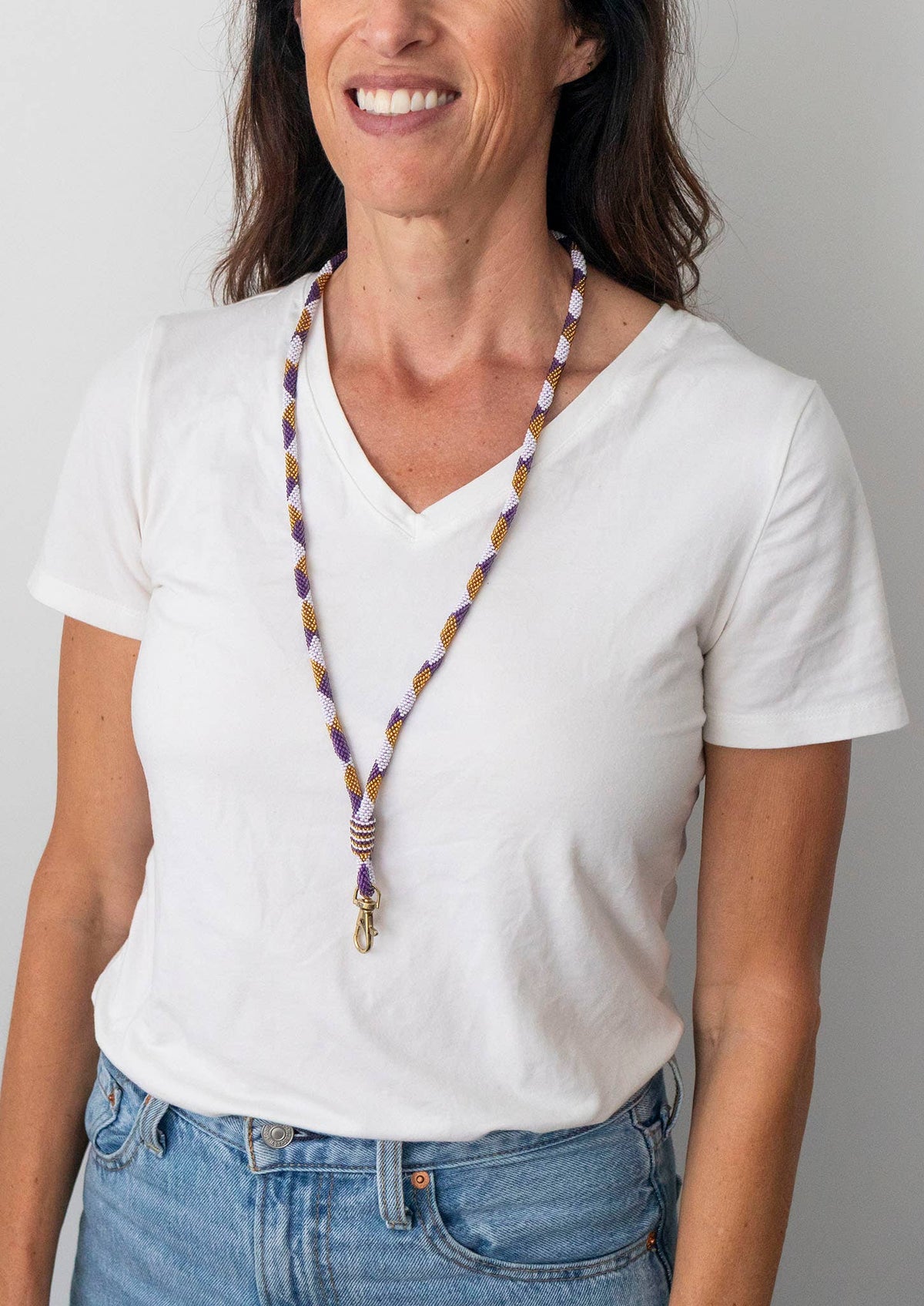 A woman wearing a white V-neck t-shirt and blue jeans, showcasing a long beaded necklace with purple, gold, and white beads and a gold clasp.