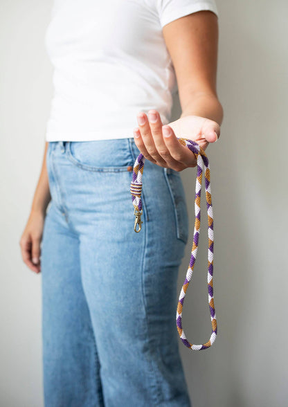 A person wearing a white t-shirt and blue jeans holds a braided rope dog leash with purple, white, and gold stripes and a silver metal clasp against a plain light gray background.