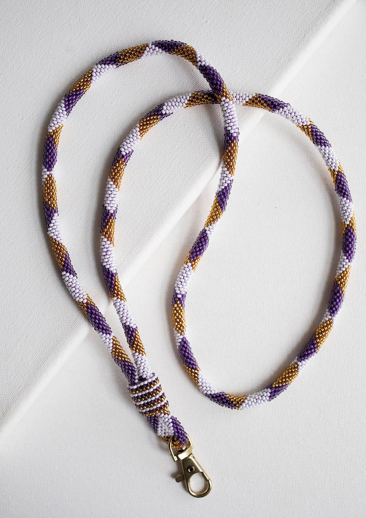 A braided rope lanyard with a gold-toned lobster clasp, featuring a pattern of purple, white, and gold twisted strands, laid out on a white background.