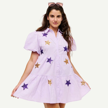 A woman modeling a light lavender short-sleeve button-up dress with purple and gold sequined star patches, set against a plain off-white background.