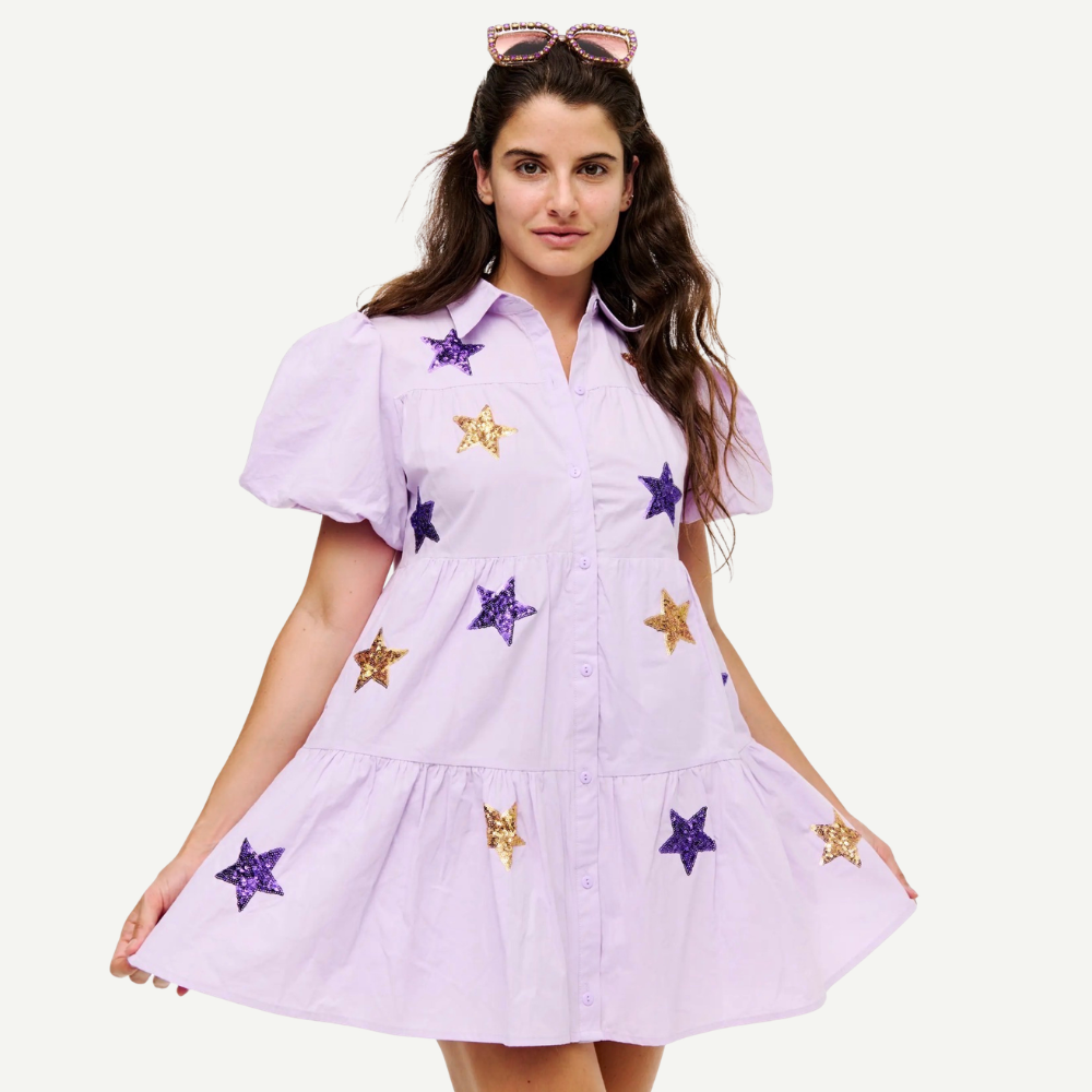 A woman modeling a light lavender short-sleeve button-up dress with purple and gold sequined star patches, set against a plain off-white background.