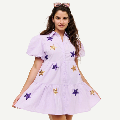 A woman modeling a light lavender short-sleeve button-up dress with purple and gold sequined star patches, set against a plain off-white background.